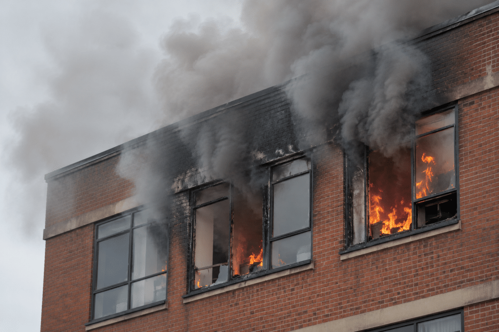 Close-up of a fire spreading through the upper floor of a brick commercial building, with orange flames visible through damaged windows, thick grey smoke billowing from the roofline and soot staining the exterior walls.