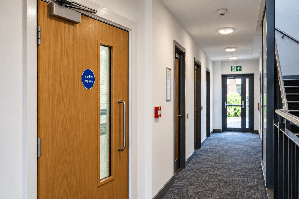 Bright, modern UK building corridor featuring a wooden fire door with a vision panel, overhead door closer and fire safety signage, with a green emergency exit sign directing occupants straight ahead towards the exit.