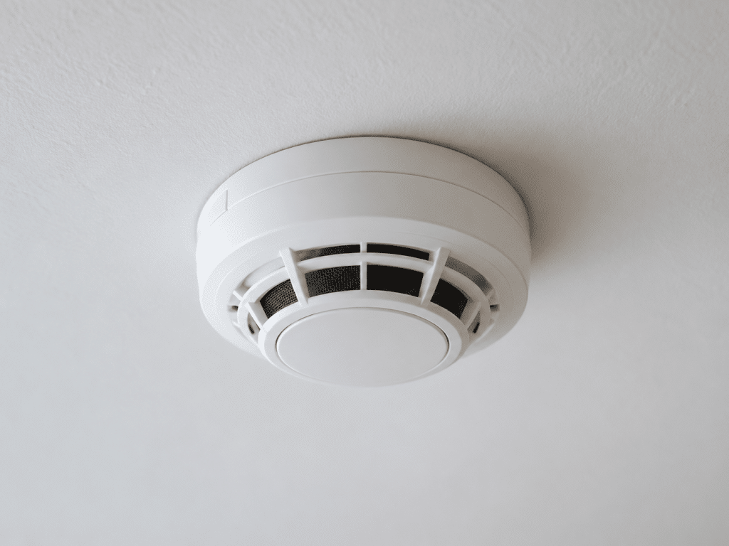 Close-up of a white ceiling-mounted smoke detector with ventilation openings, installed on a plastered ceiling to provide early fire detection and improve building fire safety.