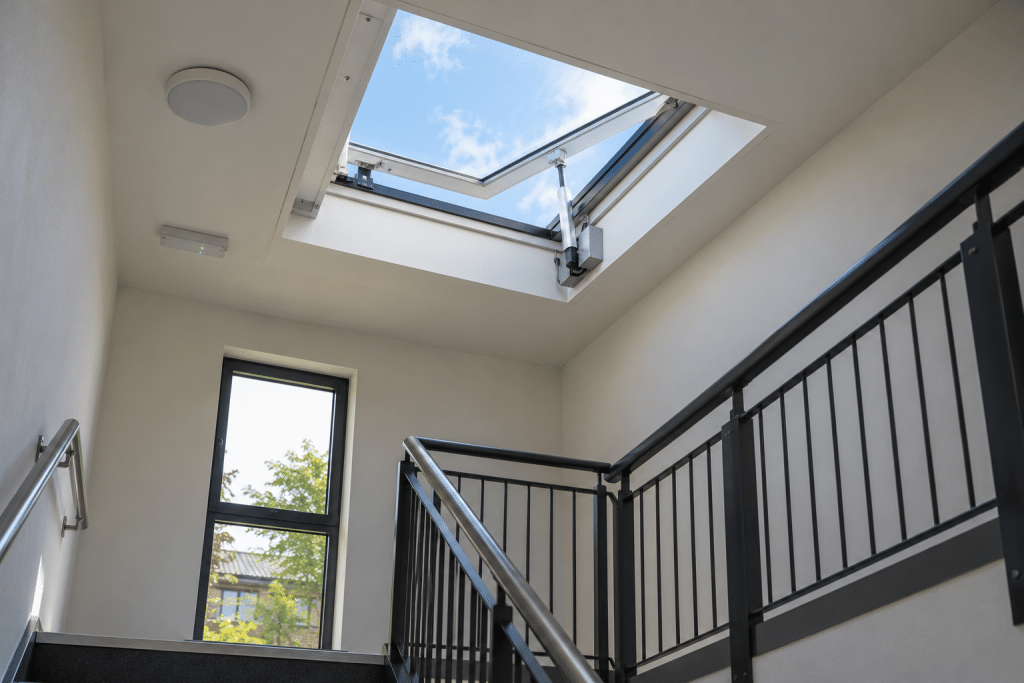 Modern UK building stairwell featuring an open roof-mounted automatic smoke vent with a visible electric actuator, allowing smoke to escape, with metal railings and natural daylight entering through the rooflight.