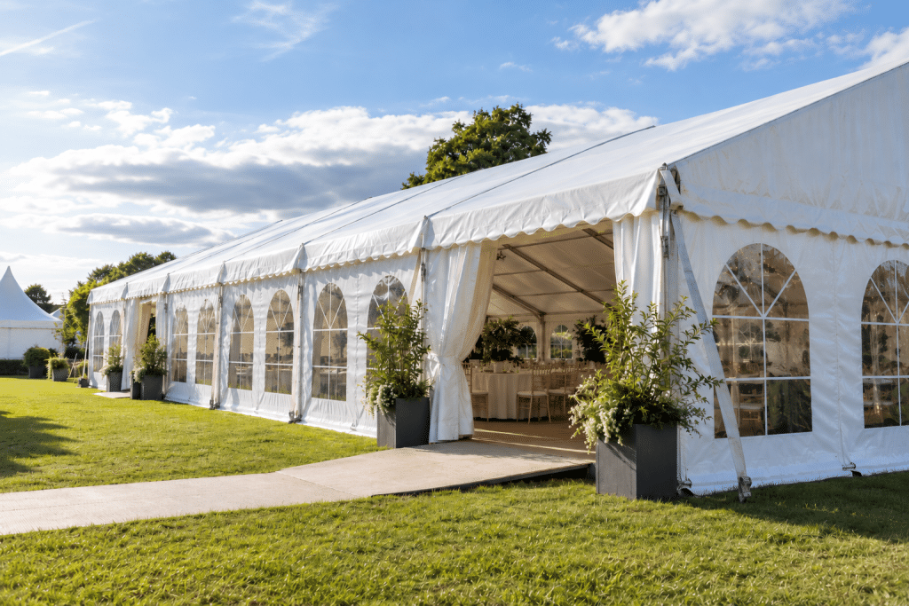 Large white event marquee set on a grassy lawn, featuring arched windows, an open entrance, a wooden walkway and decorative planters, illustrating a temporary structure used for outdoor events.