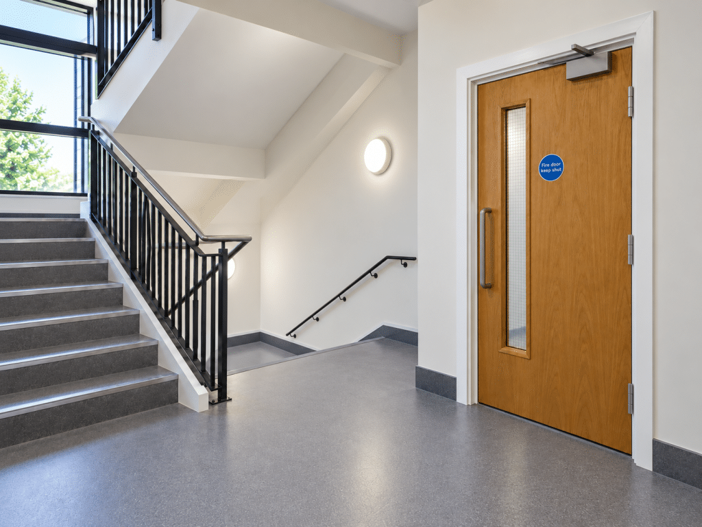 Modern stairwell with a wooden fire door featuring a vision panel, self-closing mechanism and “Fire door keep shut” sign.