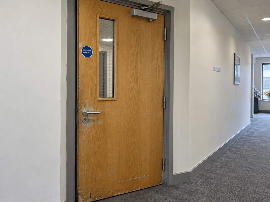 Worn wooden fire door in an office corridor, showing surface damage, a vision panel, self-closing mechanism and “Fire door keep shut” sign.