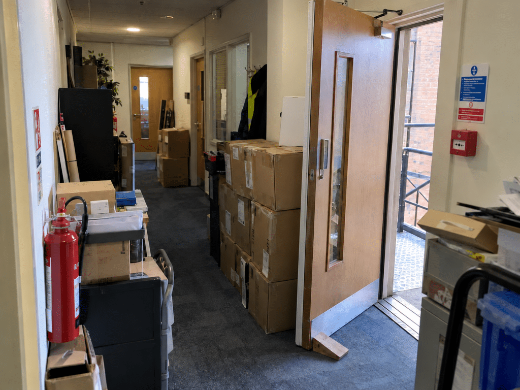 Cluttered office corridor with stacked boxes obstructing an open exit door, alongside a fire extinguisher and manual alarm call point.