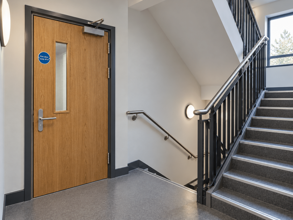Wooden fire door with a self-closing mechanism, vision panel and “Fire door keep shut” sign beside a protected stairwell.