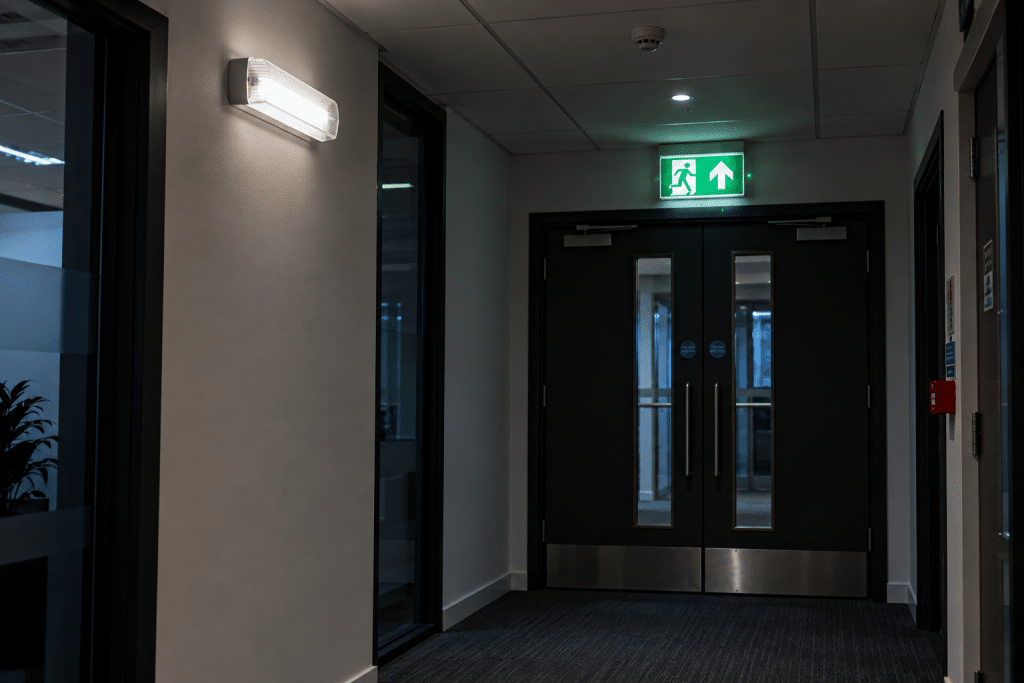 Emergency lighting illuminating a dim office corridor, with a clearly marked fire exit sign above double exit doors.