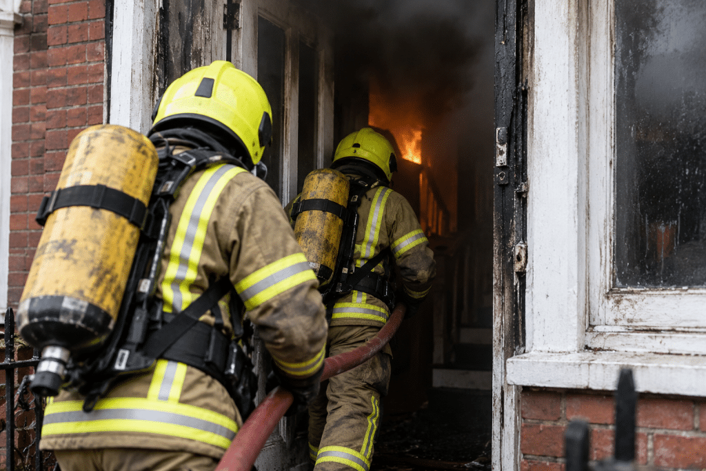Alt text: Firefighter in full protective gear climbs into a smoke-filled building during an active fire, with bright flames and thick smoke visible around the entrance.
