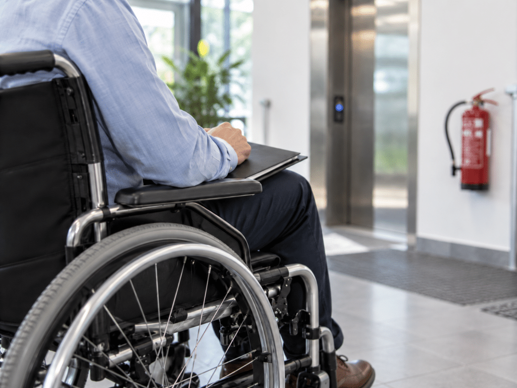 Wheelchair user viewed from behind in a modern office lobby, illustrating accessible emergency evacuation planning and Personal Emergency Evacuation Plans (PEEPs).