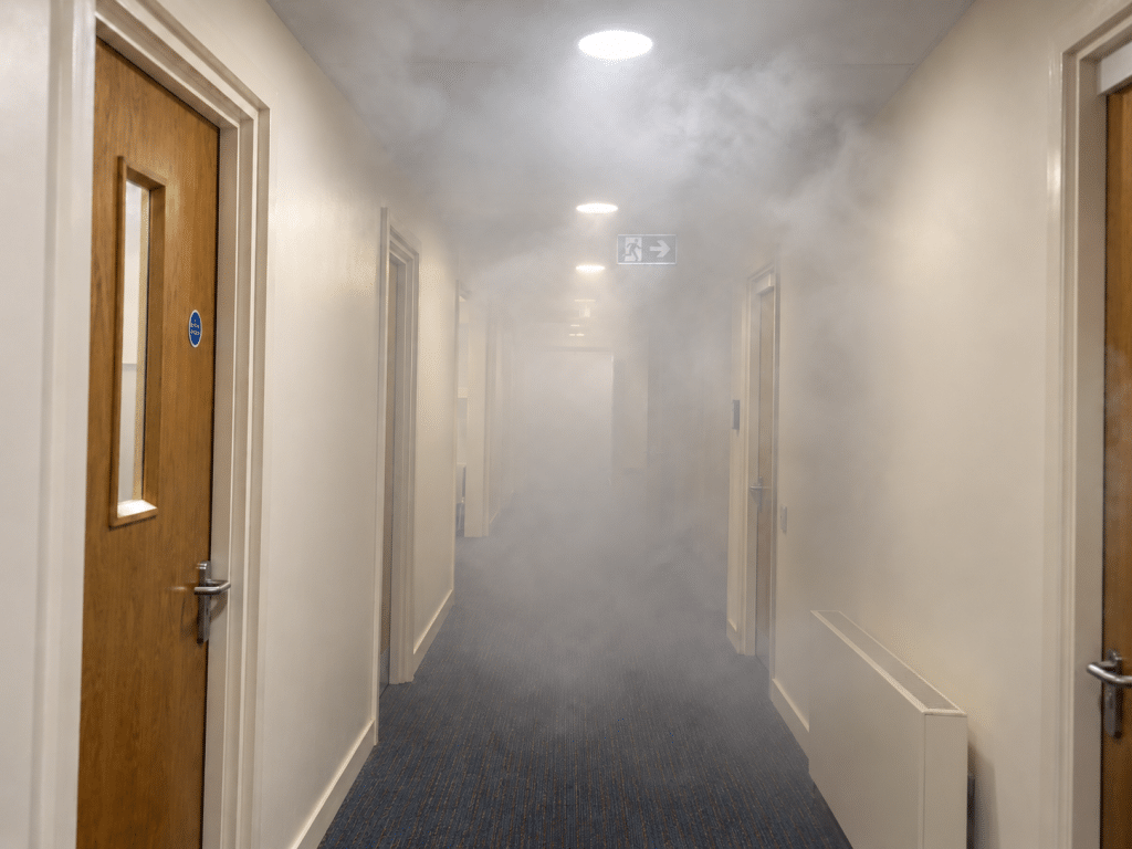 Smoke-filled commercial corridor with reduced visibility, closed fire doors and an illuminated emergency exit sign.