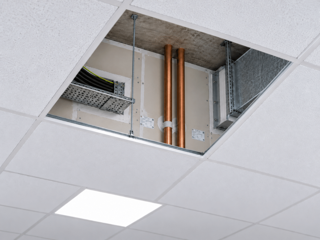 Suspended ceiling with one panel removed, revealing cables, copper pipes, ductwork and fire-stopping within the ceiling void above.