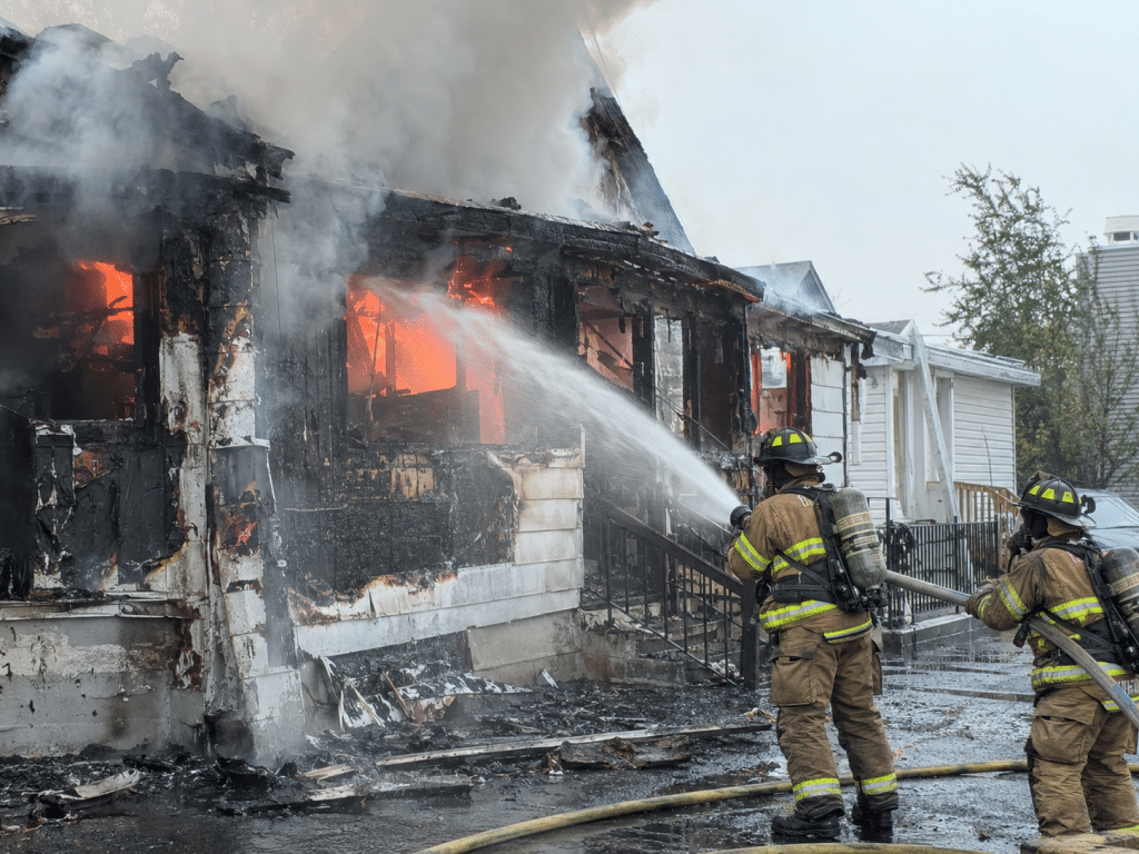 Firefighters using a high-pressure hose to tackle flames and heavy smoke at a severely fire-damaged residential building.