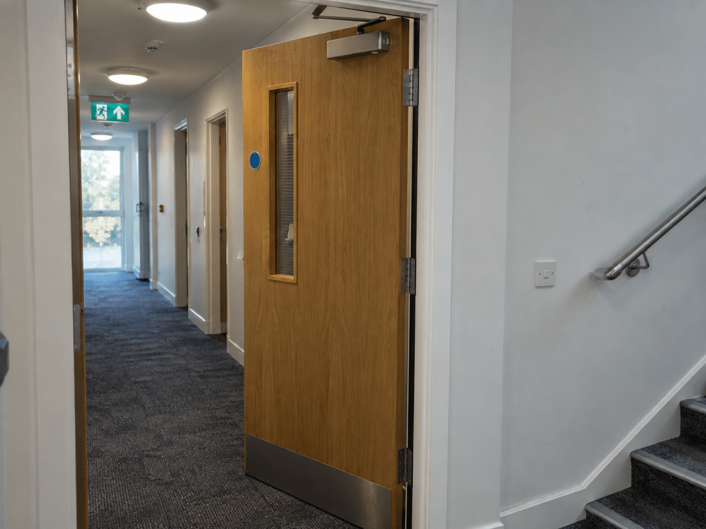 Open timber fire door with an overhead self-closing mechanism in a commercial corridor, illustrating the risk when a fire door fails to close fully.