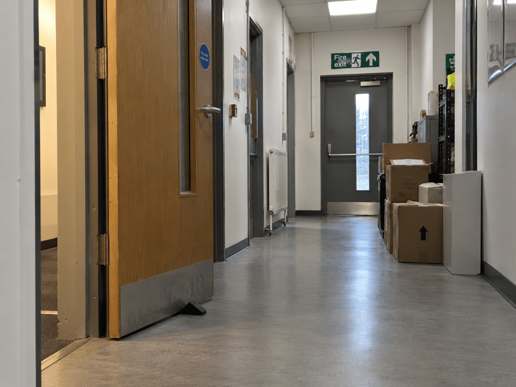Commercial corridor with a fire door wedged open and cardboard boxes stored along the escape route, illustrating temporary fixes that can become permanent fire hazards.