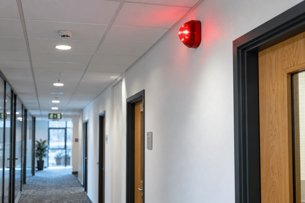 Wall-mounted visual fire alarm beacon flashing near ceiling level in a modern workplace corridor with a fire exit visible in the distance.