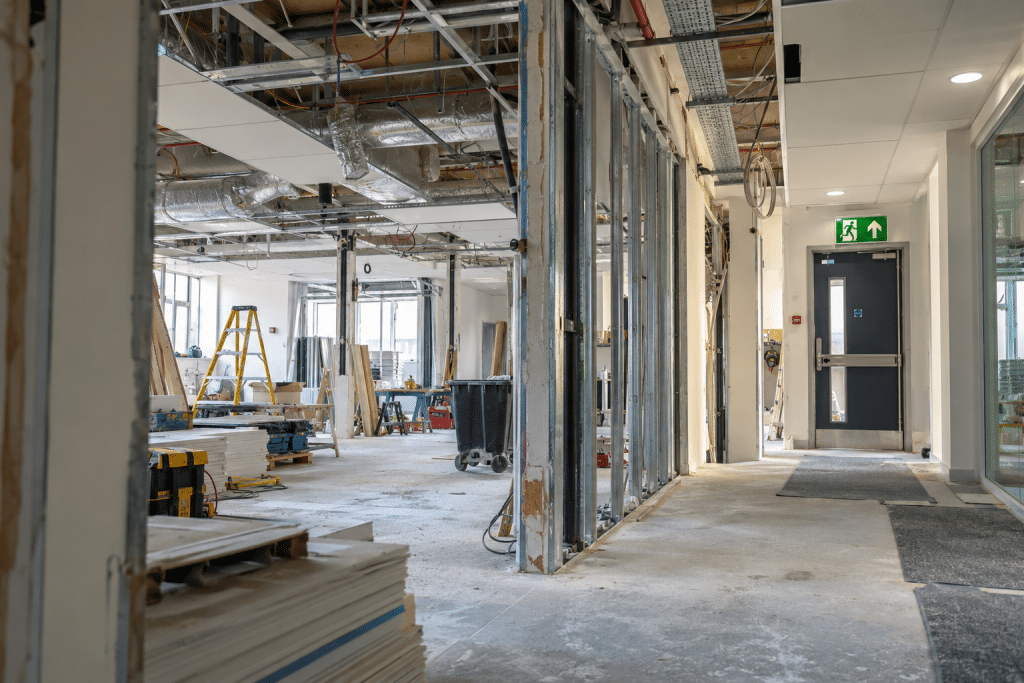 Commercial building undergoing major internal renovation, with exposed walls and ceiling services beside a finished corridor and clearly marked fire exit.