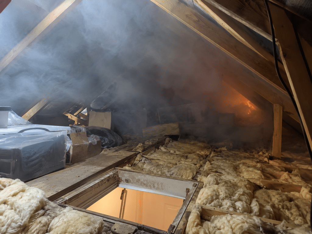 Smoke filling a residential loft space with exposed timber rafters, insulation and stored items, showing how fire and smoke can spread through an attic.